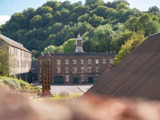 A brown brick building with lots of windows and a clocktower is visible in the distance across a car park and some picnic benches. The corner of another building is visible in the foreground.