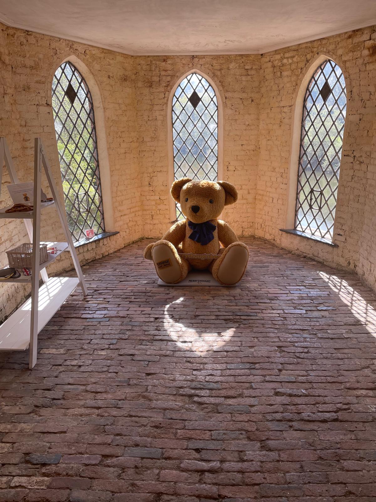 A life-sized teddy bear with  a dark blue bow around its neck sat on a cobbled floor with windows behind it.