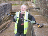 A man, standing, smiling towards the camera, wearing a high-vis jacket and a badge saying "Volunteer", standing outdoors and holding a broom, with dusty brick walls beside and behind him.