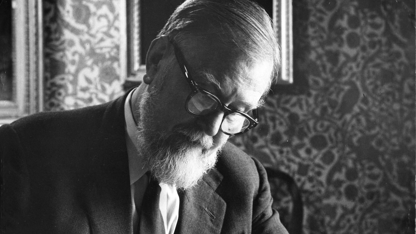 A black and white photograph of a man with glasses and white hair, wearing a suit, looking at an album of prints.