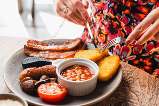 A woman is using a knife and fork to cut a hash brown on her breakfast plate. There is also bacon, sausage, baked beans, tomato, mushrooms and black pudding on the plate.