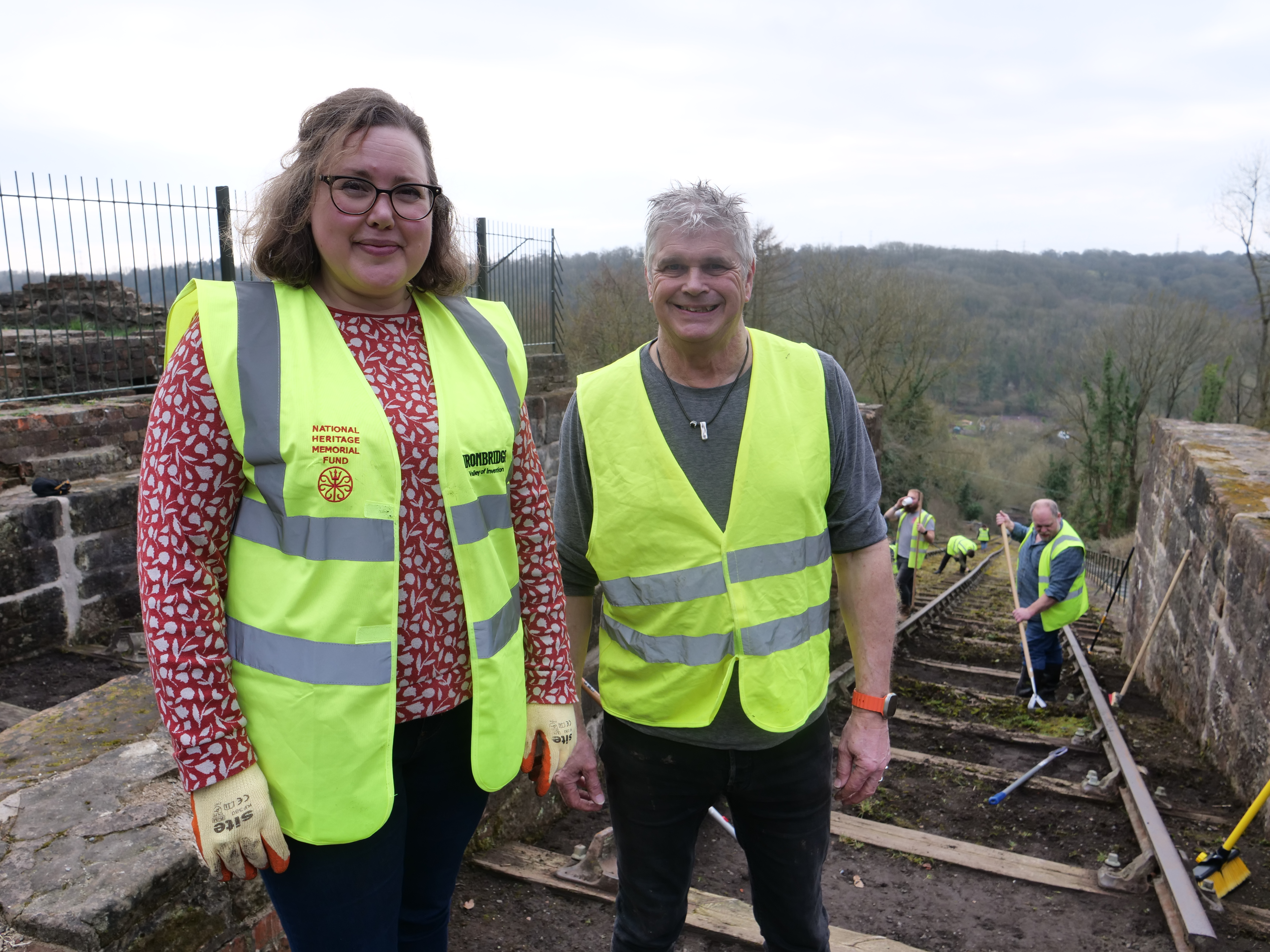 Smiling woman and man wearing high visibility jackets in front of a railway track being cleaned by another man with a broom 