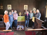 A group of seven women wearing badges saying "Volunteer", accompanied by one woman wearing Victorian costume and one woman wearing a badge saying "Staff", all standing in a wooden chapel behind a rag rug depicting a cross. They are all smiling.